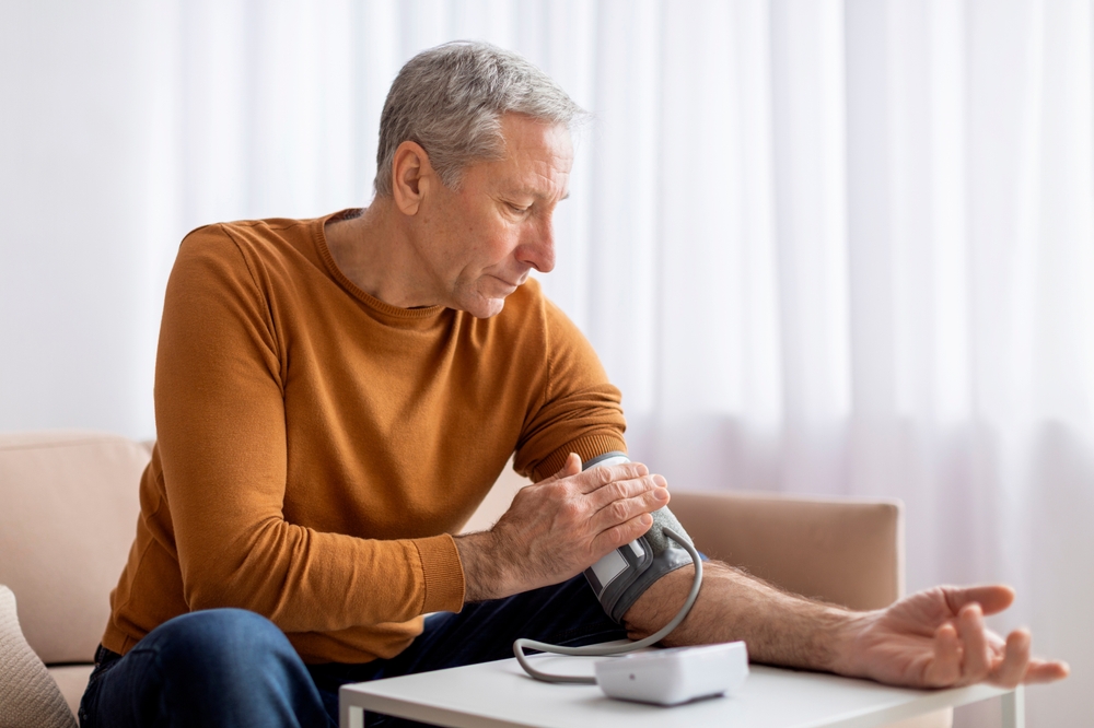Determined senior man in casual outfit sitting on couch at home, checking blood pressure on his own. Pensioner using modern tonometer, experiencing hypertension
