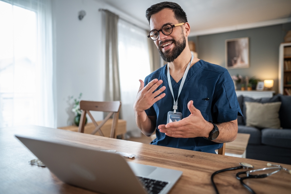 Young male doctor in scrubs conducting an online consultation on his laptop, actively gesturing while providing membership-based primary care